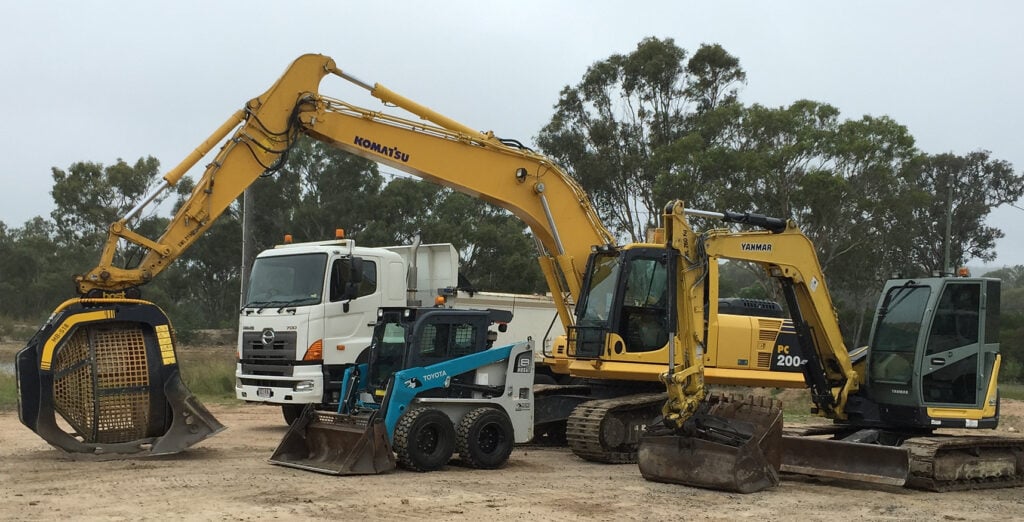 diverse excavation fleet featuring multiple heavy machinery, including Komatsu and Yanmar excavators, a Toyota skid-steer loader, and a Hino dump truck, parked at a construction site with trees in the background.